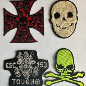 Op de afbeelding: Vier geborduurde patches met schedelontwerpen. De eerste patch is een rood kruis met zwarte schedels. De tweede patch is een witte schedel met zwarte ogen en een glimlach. De derde patch is een zwarte patch met een zilveren schedel en de tekst "ESC 153 TOUGH O". De vierde patch is een groene schedel en gekruiste beenderen.