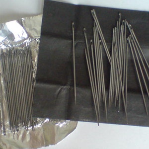 May include: Two packages of silver sewing needles. One package is wrapped in aluminum foil and the other is wrapped in black paper.