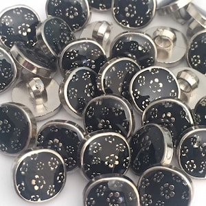May include: A pile of black buttons with silver trim and a floral design. The buttons are round and have a flat back.