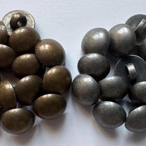 Può includere: Una collezione di bottoni rotondi a forma di cupola disposti in due gruppi distinti. Il gruppo a sinistra è composto da bottoni con una finitura metallica color bronzo scuro, mentre il gruppo a destra presenta bottoni con una finitura argentata.