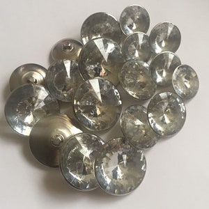 May include: A group of clear crystal buttons with silver backs. The buttons are arranged in a loose pile on a white surface.
