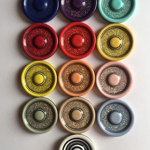 May include: Twelve round, decorative buttons with a variety of colors and glitter finishes. The buttons have a raised center with a black ring around the edge. The buttons are arranged in three rows of four.