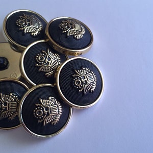 Pode incluir: Seis botões pretos com acabamento dourado e um design de águia. Os botões estão dispostos em círculo.