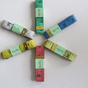May include: Six different colored measuring tapes arranged in a star shape. The tapes are all 60 inches or 150 centimeters long. The tapes are yellow, white, blue, green, pink, and red.