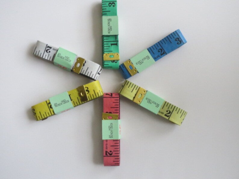 May include: Six different colored measuring tapes arranged in a star shape. The tapes are all 60 inches or 150 centimeters long. The tapes are yellow, white, blue, green, pink, and red.