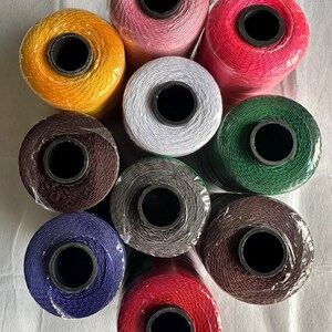 May include: A collection of colorful spools of thread in various shades, including yellow, pink, red, white, green, brown, blue, and gray. Each spool is tightly wound and wrapped in clear plastic, showcasing the vibrant colors of the thread.