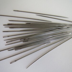 May include: A collection of silver sewing needles arranged in a fan shape on a white surface.