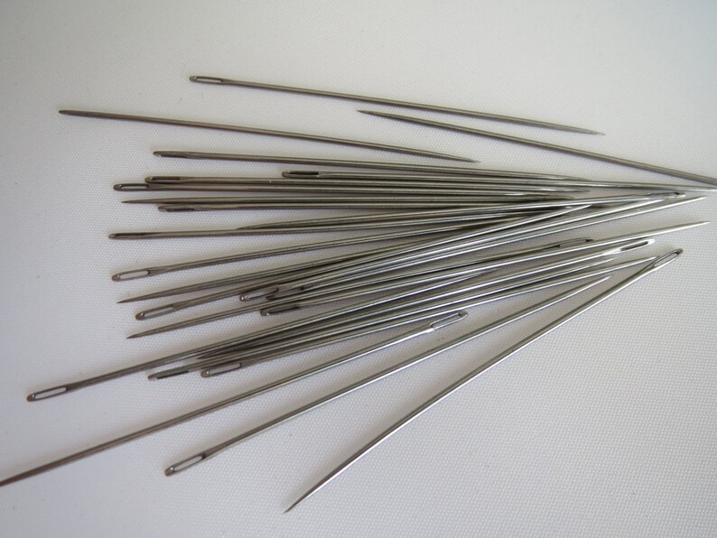 May include: A collection of silver sewing needles arranged in a fan shape on a white surface.