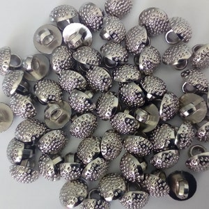 May include: A large group of silver metal buttons with a textured dome shape. The buttons have a shiny finish and are arranged in a random pattern.