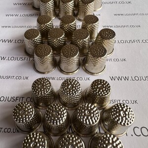 May include: A collection of 25 silver thimbles with a textured surface. The thimbles are arranged in a circular pattern on a white surface.