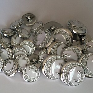 May include: A pile of white and silver round buttons with a decorative swirl design. The buttons have a raised edge and a shiny finish.