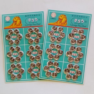 May include: Two packs of silver spring press studs, arranged in a floral pattern on teal cards. Each card features a golden lion logo and the text "999 RUST PROOF Spring Press Stud".