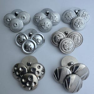 May include: A collection of ten silver-toned buttons with various designs. Some buttons have a raised design, while others have a textured surface. The buttons are all approximately the same size.