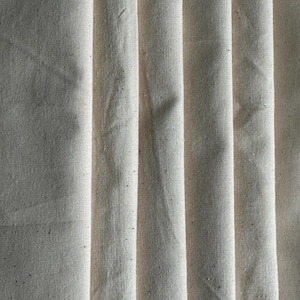 Puede incluir: Primer plano de tela blanquecina con pliegues verticales. La tela tiene un aspecto texturizado con sutiles variaciones de tono y pequeñas motas oscuras. Los pliegues crean sombras y reflejos, añadiendo profundidad a la imagen. La tela parece ser un material natural.