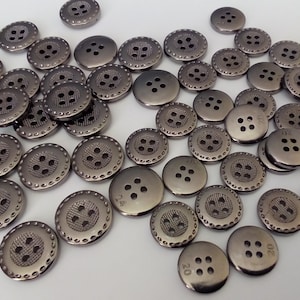 May include: A pile of silver metal buttons with four holes each. The buttons have a decorative, textured design around the edge.