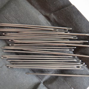 Może przedstawiać: Zbliżenie na grupę srebrnych igieł do szycia ułożonych w stos na czarnej powierzchni.