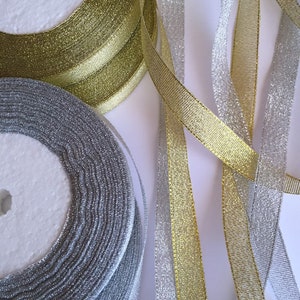 May include: Close-up of spools of gold and silver metallic ribbon. The ribbon is unwound in sections, revealing its shiny texture. The spools are white, and the background is a plain white surface. The ribbons are ideal for crafts and gift wrapping.