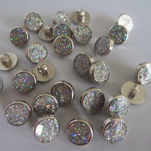 May include: A collection of silver-toned buttons with a sparkly iridescent finish. The buttons are round and have a flat back.