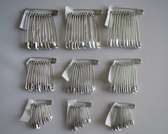May include: A set of silver safety pins, arranged in rows on a white surface. The pins are of various sizes, with some being larger than others.