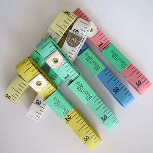 May include: A set of five colorful measuring tapes, each with a different color and markings. The tapes are made of fabric and have a metal end cap. The tapes are all in inches and centimeters. The tapes are arranged in a star shape.
