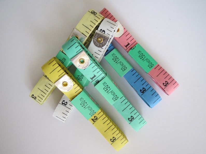 May include: A set of five colorful measuring tapes, each with a different color and markings. The tapes are made of fabric and have a metal end cap. The tapes are all in inches and centimeters. The tapes are arranged in a star shape.