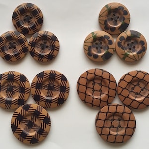 May include: Nine wooden buttons with different patterns. The buttons are brown with black designs. Some buttons have a checkered pattern, some have a floral pattern, and some have a net pattern.