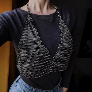May include: A silver chainmail top with a V-neckline. The top is worn over a black long-sleeved shirt and blue jeans.
