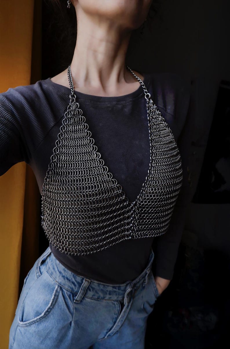 May include: A silver chainmail top with a V-neckline. The top is worn over a black long-sleeved shirt and blue jeans.