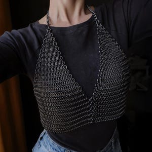 May include: A silver chainmail top with a V-neckline. The top is worn over a black shirt and blue jeans.