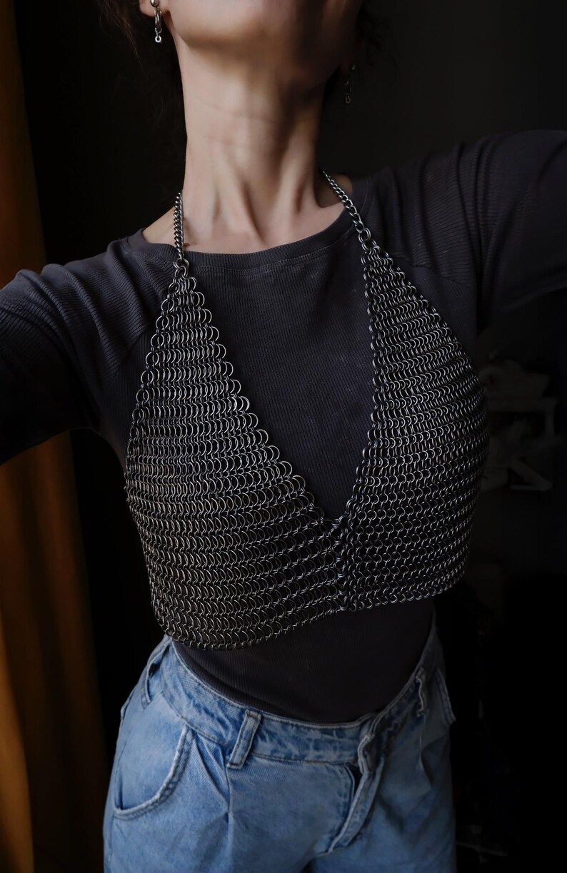 May include: A silver chainmail top with a V-neckline. The top is worn over a black shirt and blue jeans.