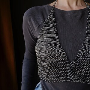 May include: A silver chainmail top with a halter neckline. The top is made of small, interconnected metal rings. The top is worn over a black long-sleeved shirt and blue jeans.