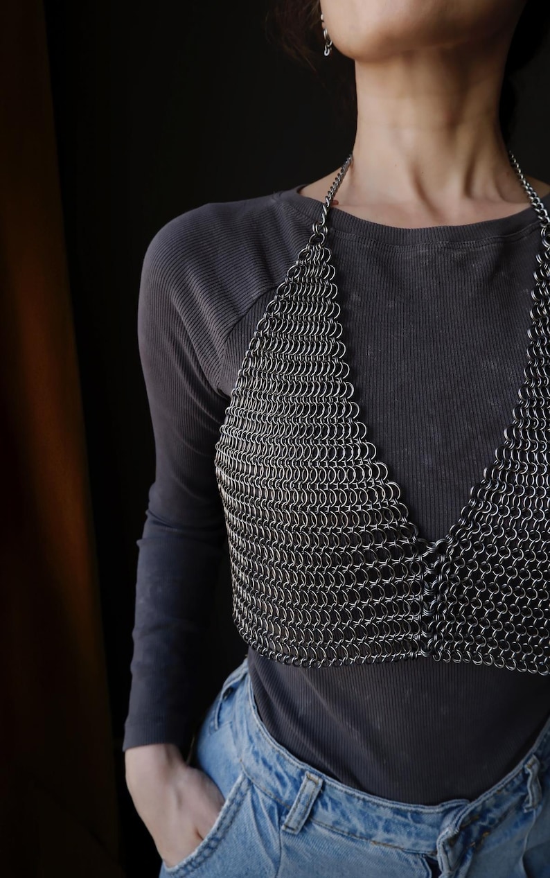 May include: A silver chainmail top with a halter neckline. The top is made of small, interconnected metal rings. The top is worn over a black long-sleeved shirt and blue jeans.