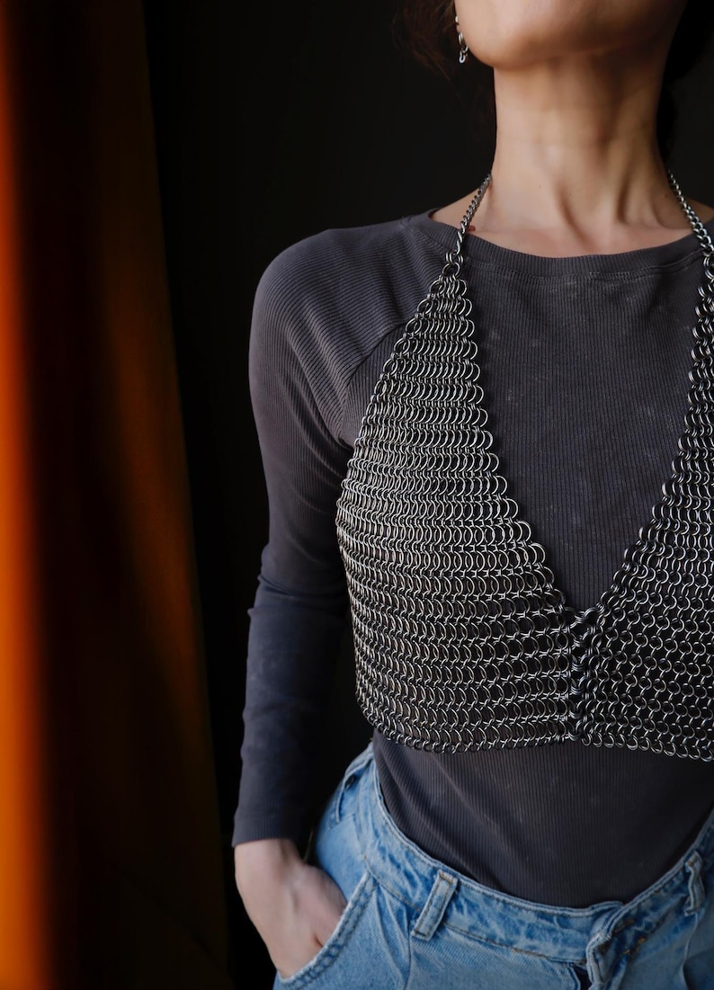 May include: A silver chainmail top with a halter neckline. The top is made of small, interlocking metal rings. The top is worn over a gray long-sleeved shirt and blue jeans.