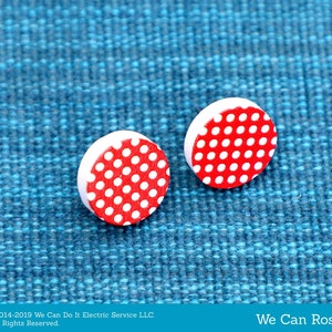 Pode incluir: Dois brincos redondos de madeira com um padrão de bolinhas vermelhas e brancas. Os brincos estão sobre uma superfície azul texturizada.