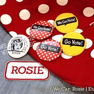 Op de afbeelding: Vier rode, witte en gele knoopsluitingen met de tekst "We Can Vote!" en "Go Vote!" op een rode achtergrond met witte stippen. Een witte knoopsluiting met de tekst "Westinghouse Electric Service" is ook zichtbaar.