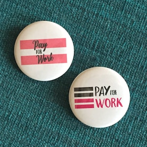 May include: Two white button pins with black text that reads "Pay for Work". One pin has pink stripes and the other has black stripes.