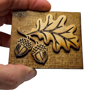 May include: A rectangular wooden plaque featuring a laser-engraved design of an oak leaf and two acorns. The wood has a medium brown tone with a visible grain pattern, and the engraved lines are dark brown, creating a high-contrast, detailed image.