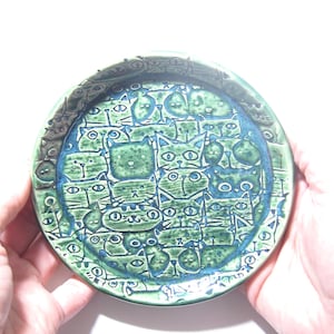 Może przedstawiać: Okrągły ceramiczny talerz z gęstym, tłoczonym wzorem przedstawiającym liczne kocie pyszczki w odcieniach głębokiego błękitu i leśnej zieleni. Wzór pokrywa całą powierzchnię, w tym podniesiony brzeg, z precyzyjnymi liniami definiującymi każdego kota.