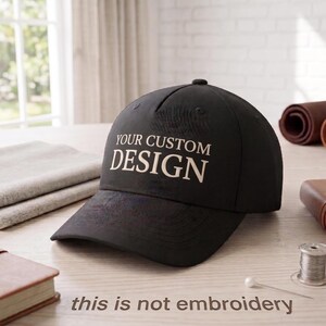 May include: A black baseball cap with the text "YOUR CUSTOM DESIGN" in white. The cap is on a light-colored surface, with a needle and thread visible. The phrase "this is not embroidery" is at the bottom.