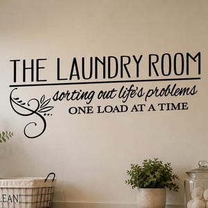 Op de afbeelding: Een zwarte muursticker met de tekst THE LAUNDRY ROOM, sorting out life's problems en ONE LOAD AT A TIME, vergezeld van een decoratief krulontwerp. De belettering is aangebracht op een effen muur boven een draadmand en een glazen pot.