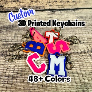 May include: A collection of custom 3D printed keychains featuring various letters in vibrant colors including pink, blue, orange, and white. Each keychain is attached to a silver-toned metal lobster clasp. The text Custom 3D Printed Keychains and 48+ Colors is visible.