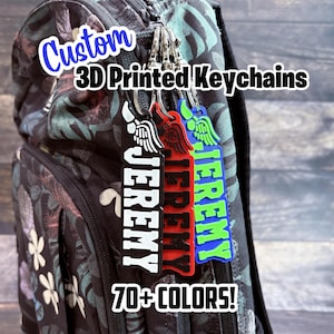 May include: Custom 3D printed keychains in black, red, and blue, each with the name "Jeremy" in white. The keychains feature wing designs and are attached to a backpack. The image includes the text "70+ COLORS!"