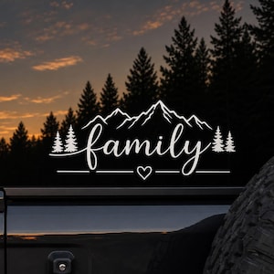Op de afbeelding: Een witte vinyl sticker op een voertuigruit met een silhouet van een bergketen boven het woord family in een cursief lettertype, geflankeerd door dennenbomen aan beide kanten, met een klein hartje onder de tekst.