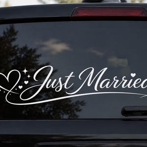 Könnte beinhalten: Ein weißer Vinyl-Aufkleber auf einer dunklen Autoscheibe mit dem geschriebenen Text Just Married. Das Design enthält eine Herz-Kontur auf der linken Seite, kleine dekorative Sterne und drei winzige Herzen, die vom Haupt-Herz ausgehen.