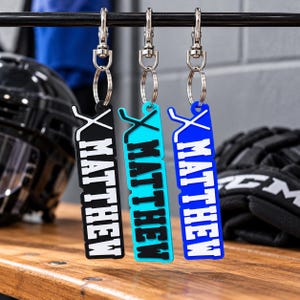 May include: Three rectangular keychains hanging from a metal bar, each featuring a crossed hockey stick icon at the top and the name MATTHEW printed in bold block letters. The keychains are black with white text, turquoise with black text, and blue with white text.