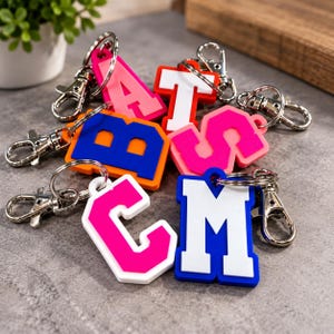 May include: A collection of letter keychains featuring varsity-style typography in vibrant colours including hot pink, orange, blue, and white. Each keychain is attached to a silver-toned metal lobster clasp and split ring, showcasing letters A, B, C, M, S, and T.