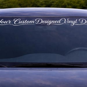 Puede incluir: Una calcomanía de vinilo blanca con el texto "Your Custom Designed Vinyl Decal" en una fuente elegante, aplicada al parabrisas de un coche azul oscuro. La calcomanía está colocada en la parte superior del parabrisas.