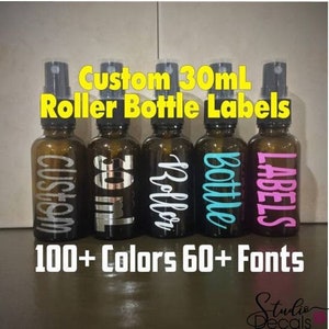 Könnte beinhalten: Fünf bernsteinfarbene Glas-Rollflaschen mit individuellen Etiketten. Die Etiketten sind weiß mit schwarzem Text. Der Text auf den Etiketten lautet "Custom", "30ml", "Roller", "Bottle" und "Labels". Die Flaschen sind von links nach rechts in einer Reihe angeordnet. Der Text "100+ Farben 60+ Schriftarten" befindet sich unter den Flaschen.
