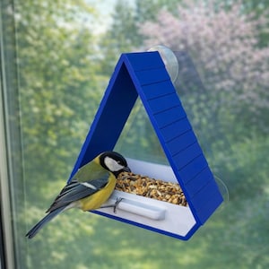 Puede incluir: Un comedero triangular azul para pájaros fijado a una ventana con una ventosa. Un pequeño pájaro con plumas amarillas, negras y blancas se posa en el comedero, comiendo semillas. El comedero tiene una bandeja blanca y está lleno de alpiste.