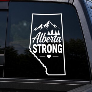 May include: A white vinyl decal applied to a dark vehicle window, featuring the outline of the province of Alberta. Inside the shape are mountain peaks, pine trees, the text Alberta STRONG, and a small heart symbol centered below the text.